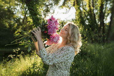 Blond woman smelling peony flower while standing at meadow - LLUF00015