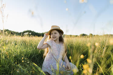 Smiling girl holding hat while kneeling at meadow - LLUF00023