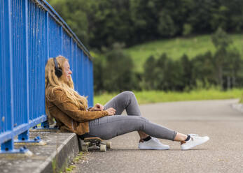 Young woman listening to music on headphones while sitting on skateboard and leaning on railing of Remsbrucke bridge - STSF03030