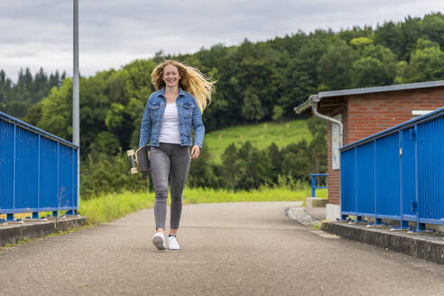 Junge Frau lächelt in die Kamera, während sie mit einem Skateboard in der Hand über die Remsbrucker Brücke läuft - STSF03031