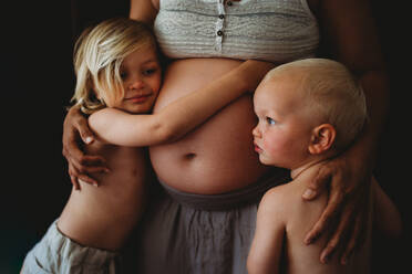 Beautiful blonde kids hugging mom's pregnant big belly at home - CAVF94889