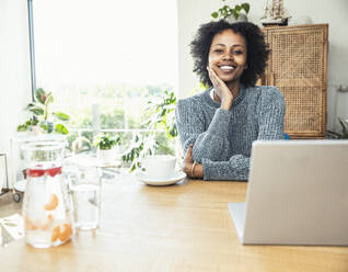 Smiling female professional with hand on chin sitting at desk - UUF24621