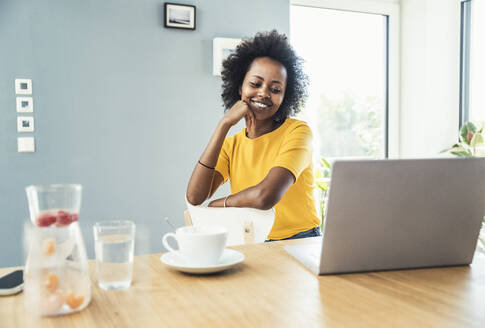 Junge Berufstätige schaut auf einen Laptop, während sie auf einem Stuhl im Heimbüro sitzt - UUF24626