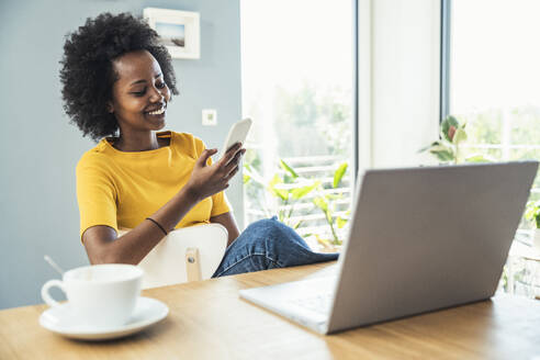 Afro-Frau, die ein Mobiltelefon benutzt, während sie zu Hause auf einem Stuhl sitzt - UUF24628