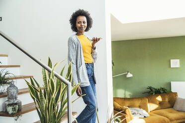 Young woman walking down on staircase while talking through phone at home - UUF24652