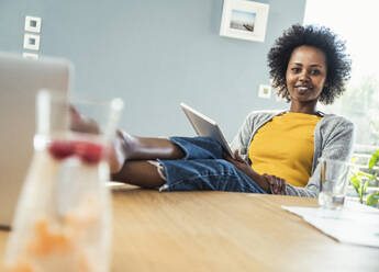 Afroamerikanische Freiberuflerin mit hochgelegten Füßen und digitalem Tablet im Heimbüro - UUF24669