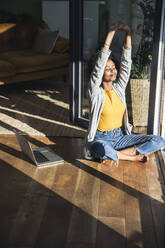 Woman with arms raised sitting cross-legged on floor at home - UUF24682