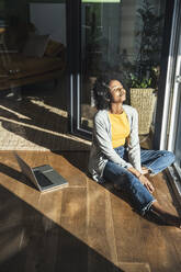 Junge Frau mit geschlossenen Augen sitzt zu Hause im Sonnenlicht - UUF24684