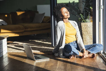 Woman sitting cross-legged by laptop in sunlight at home - UUF24687