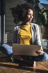 Smiling woman with laptop sitting cross-legged in sunlight at home - UUF24692