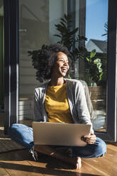 Happy woman with laptop sitting cross-legged in living room - UUF24693