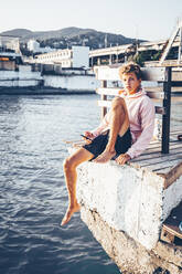 Young man holding mobile phone while sitting at pier - OMIF00007