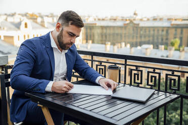Geschäftsmann schreibt auf Papier, während er auf dem Balkon sitzt - VPIF04713