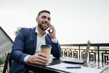 Businessman holding disposable cup while talking on smart phone in balcony - VPIF04726