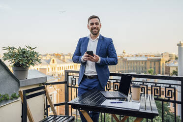 Männlicher Geschäftsmann mit Smartphone, der sich auf das Geländer eines Bürobalkons stützt - VPIF04730