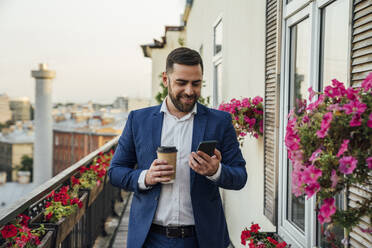 Lächelnder männlicher Fachmann, der einen Einwegbecher hält, während er ein Smartphone auf einem Balkon benutzt - VPIF04742