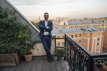 Smiling male professional with arms crossed leaning near plants in balcony - VPIF04776