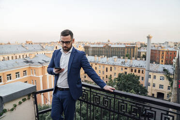 Male professional with smart phone leaning on railing in balcony - VPIF04779