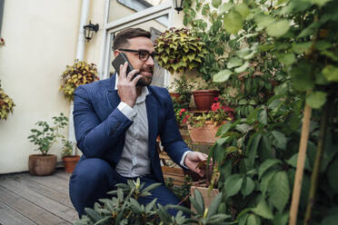 Geschäftsmann, der mit seinem Smartphone spricht, während er in der Nähe von Pflanzen auf dem Balkon hockt - VPIF04784