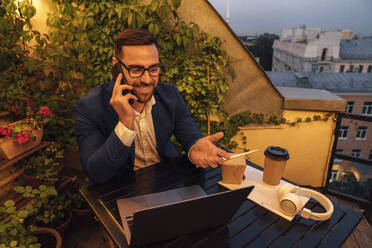 Male professional gesturing while talking on smart phone in office balcony during sunset - VPIF04799