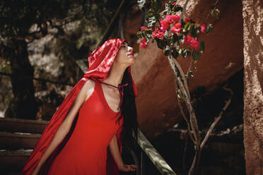 Frau in rotem Umhang riecht mit geschlossenen Augen an Blumen auf einem Baum - MRRF01501