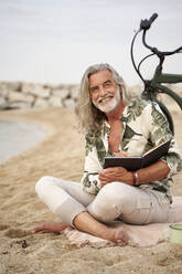 Lächelnder Mann mit Buch im Schneidersitz am Strand - VEGF04966