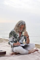 Man holding mug while writing in book at beach - VEGF04980