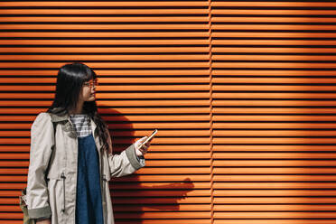 Junge Frau mit Smartphone vor einer orangefarbenen Wand - ASGF01463