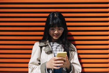Junge Frau mit Brille und Smartphone vor einer orangefarbenen Wand - ASGF01464