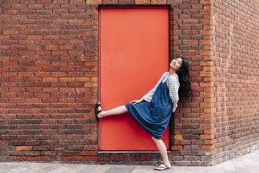 Glückliche Frau lehnt vor einer roten Tür an einer Backsteinmauer - ASGF01481