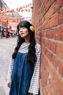 Nachdenkliche Frau hört Musik über Kopfhörer, während sie sich an eine Backsteinmauer lehnt - ASGF01483