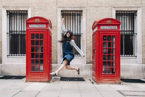 Carefree woman jumping in front of telephone booth - ASGF01488