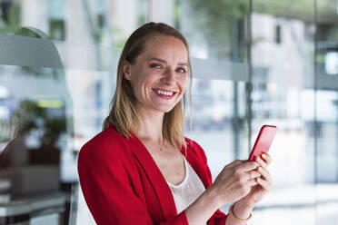 Smiling businesswoman holding mobile phone in front of glass wall - PNAF02261