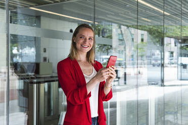 Smiling businesswoman with mobile phone standing in front of glass wall - PNAF02278