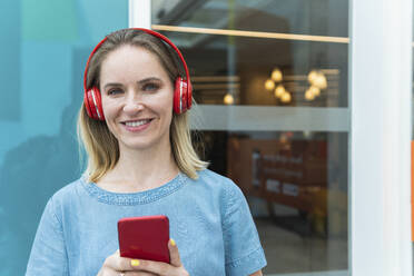 Lächelnde Geschäftsfrau mit drahtlosen Kopfhörern, die ein Mobiltelefon hält - PNAF02309