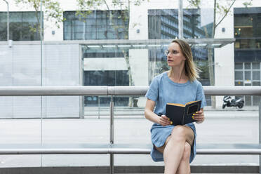 Weibliche Fachkraft mit Buch sitzt auf einer Bank vor einer Glaswand - PNAF02312
