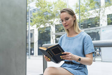 Blonde Geschäftsfrau liest ein Buch und sitzt vor einer Glaswand - PNAF02313