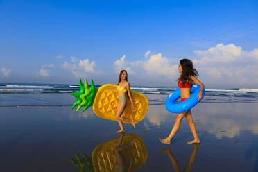 Smiling women holding swimming floats walking at beach - EAF00118