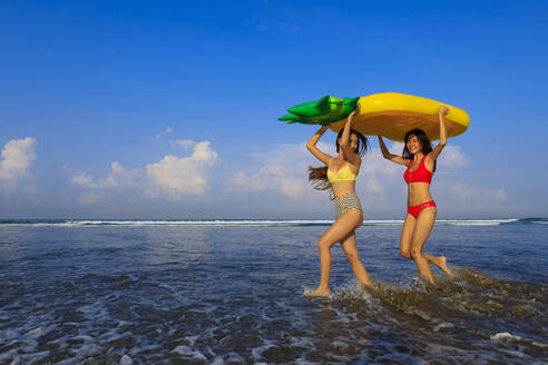 Cheerful female friends carrying pineapple float on head while enjoying at beach during vacation - EAF00122