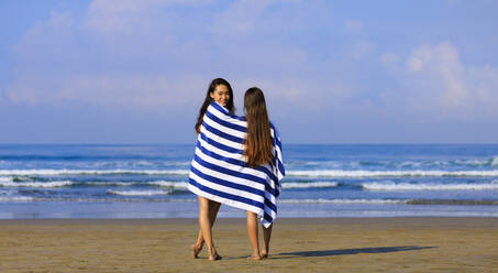 Lächelnde Frau schaut über die Schulter, während sie mit einer Freundin im Handtuch am Strand steht - EAF00125