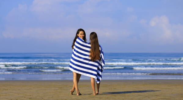 Smiling woman looking over shoulder while standing with female friend in towel at beach - EAF00125