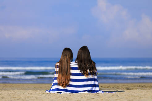 Female friends wrapped in towel sitting at beach during vacation - EAF00126