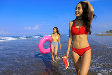 Lächelnde Frau, die eine Wassermelone in der Hand hält, während ihre Freundin mit einem Schwimmkörper an einem sonnigen Tag durchs Wasser läuft - EAF00130