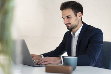 Junger Geschäftsmann tippt auf einem Laptop, während er am Schreibtisch sitzt und im Büro arbeitet - MOEF03889