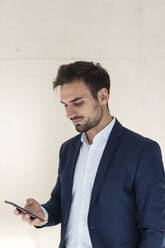 Young businessman text messaging through smart phone while standing in front of wall - MOEF03891