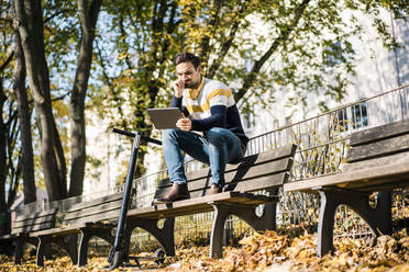 Junger Mann, der ein digitales Tablet benutzt, während er mit einem Elektroroller auf einer Parkbank in der Sonne sitzt - MOEF03892