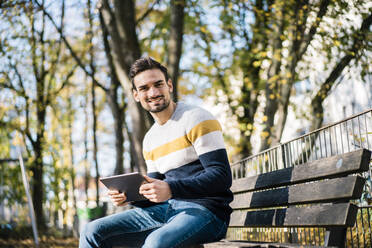 Lächelnder junger Mann hält ein digitales Tablet in der Hand, während er an einem sonnigen Tag auf einer Bank im Park sitzt - MOEF03893