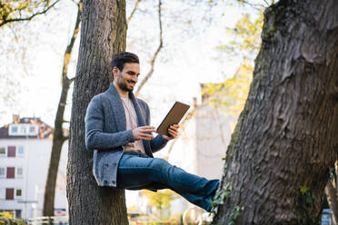 Lächelnder junger Mann sieht sich ein Video auf einem digitalen Tablet an, während er sich im Park an einen Baumstamm lehnt - MOEF03896
