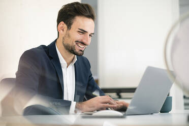 Glücklicher junger Geschäftsmann, der einen Laptop benutzt, während er am Schreibtisch im Büro sitzt - MOEF03909