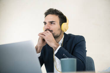 Nachdenklicher Geschäftsmann sitzt mit gefalteten Händen und hört Musik über Kopfhörer im Büro - MOEF03911